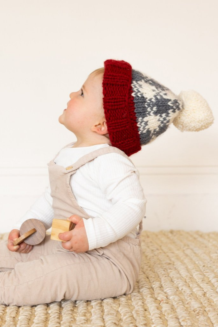 Baby boy wearing Blueberry Hill buffalo check beanie in cream, gray, and red sitting on a rug