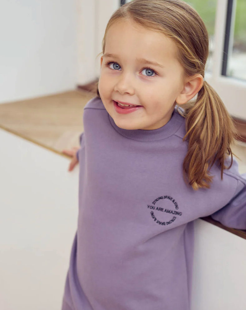 Young girl wearing a Green Cotton Group purple sweatshirt with text, sitting on a couch.