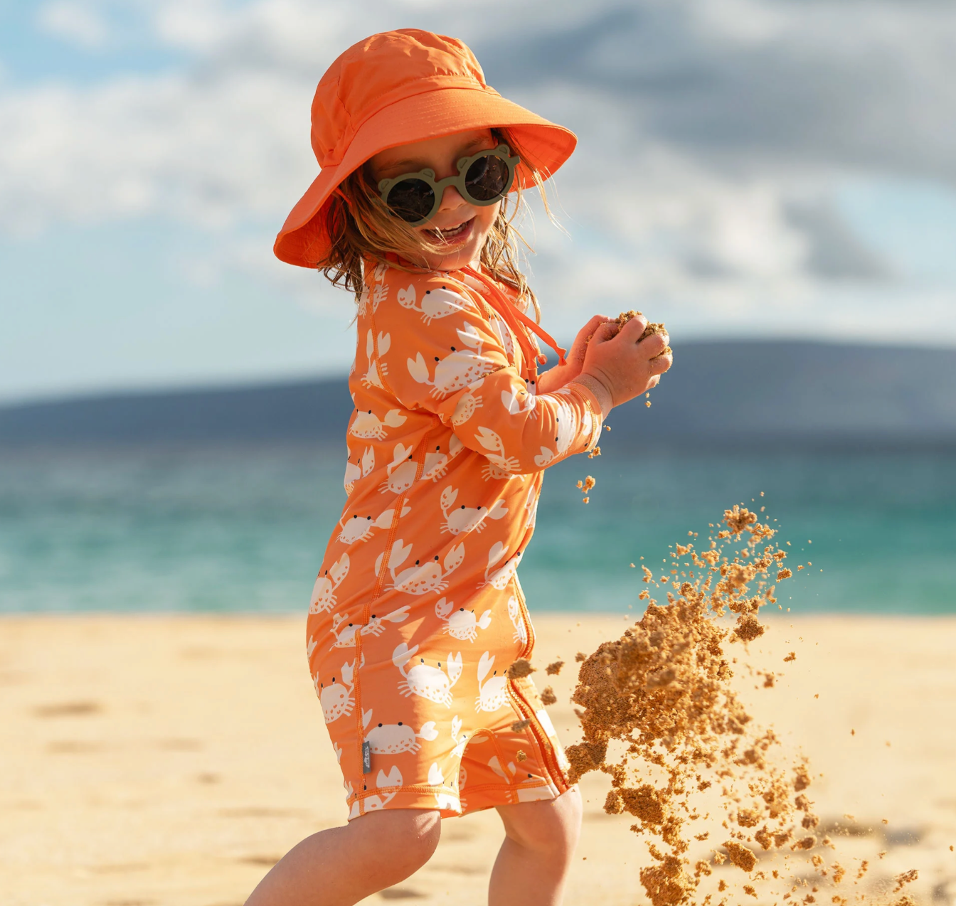 A little girl in a Jan & Jul orange bucket hat playing on the beach
