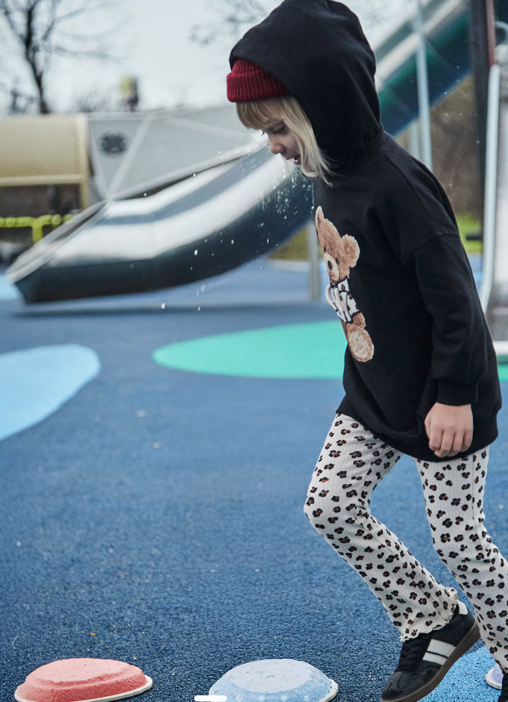 Child wearing a black hoodie with a teddy bear design and polka dot pants on a playground.