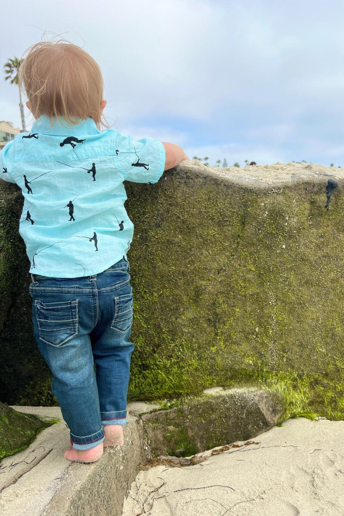 Little boy wearing Me & Henry Mark jeans on the beach leaning on a rock.