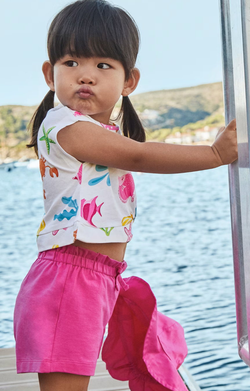 Child wearing a colorful top and pink shorts by a body of water.