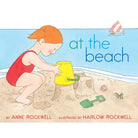 Book cover of 'At the Beach' with a child playing in the sand, a yellow bucket, and a green shovel.