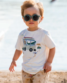Child wearing sunglasses and a graphic t-shirt with a van and palm tree design on a beach.